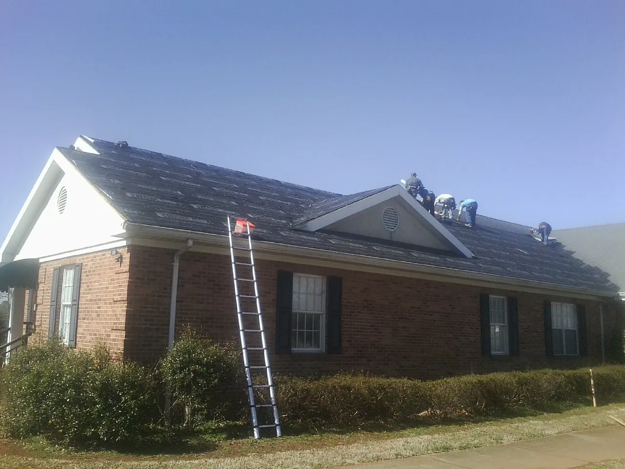 Crew tearing shingles off a brick ranch roof with a ladder set — Emergency Roof Repair work by our crew in Great Meadows