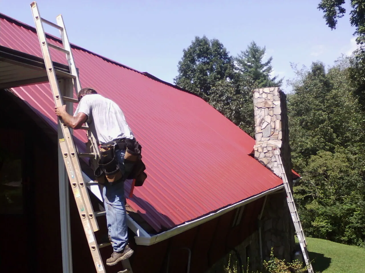 Roofer on a ladder beside a red metal roof and stone chimney — Soffit & Fascia Repair by Roofing Brothers in Great Meadows